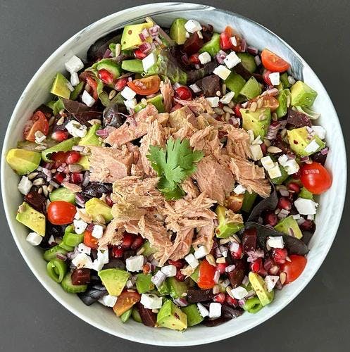 Salad bowl filled with butter lettuce, cucumbers, sugar snap peas, avocado, tomato, red onion, beets, vegan feta, and pomegranate and sunflower seeds topped with flaked tuna, JF Mustard Vinaigrette and JF Spicy Salt. 