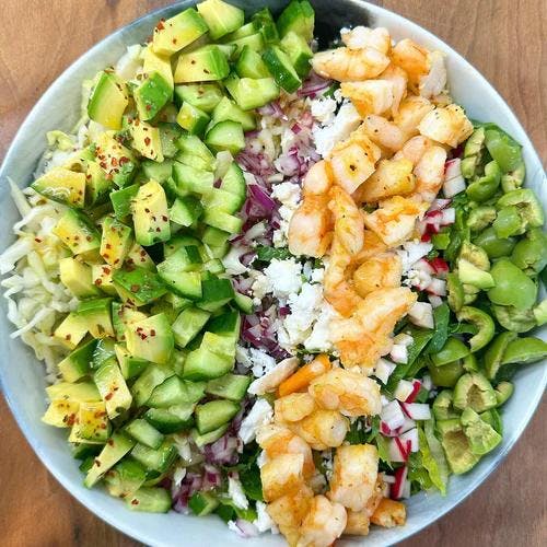 Salad with avocado, cucumber, onion, shrimp, radish and olives in white bowl on counter