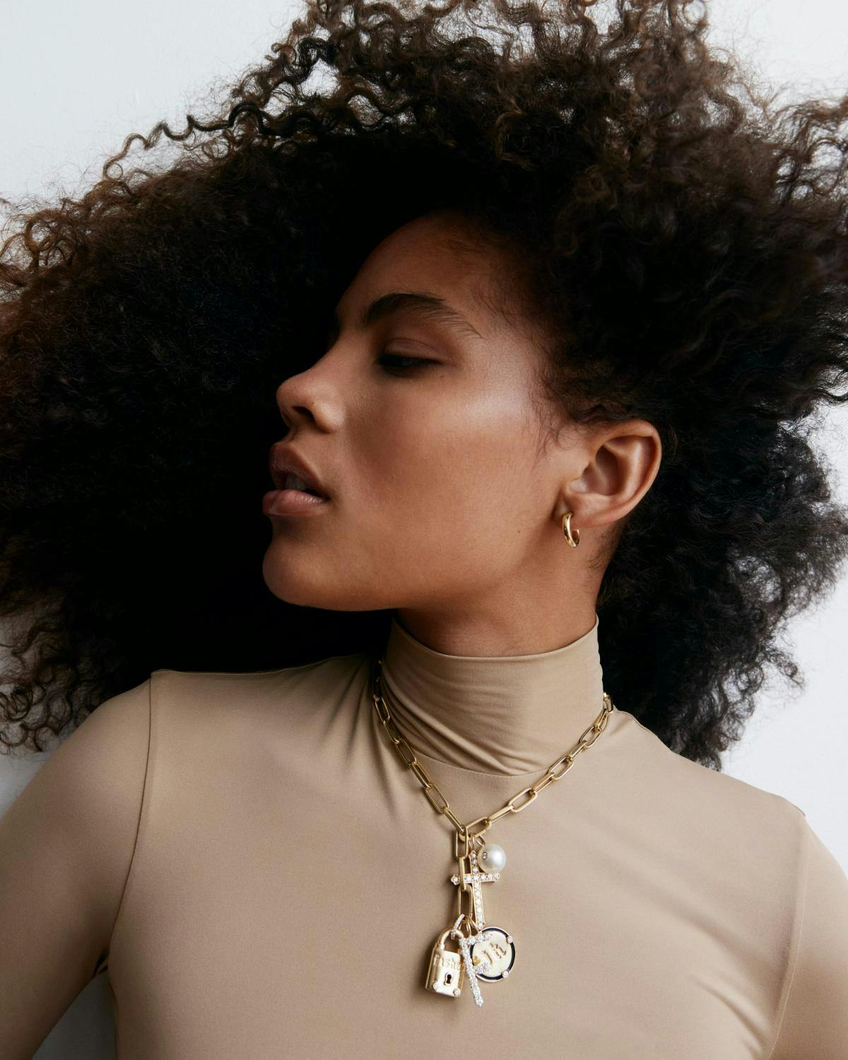 Woman in beige turtleneck with gold chain and cluster of fine gold charms including cross, lock, letter, and circle. 