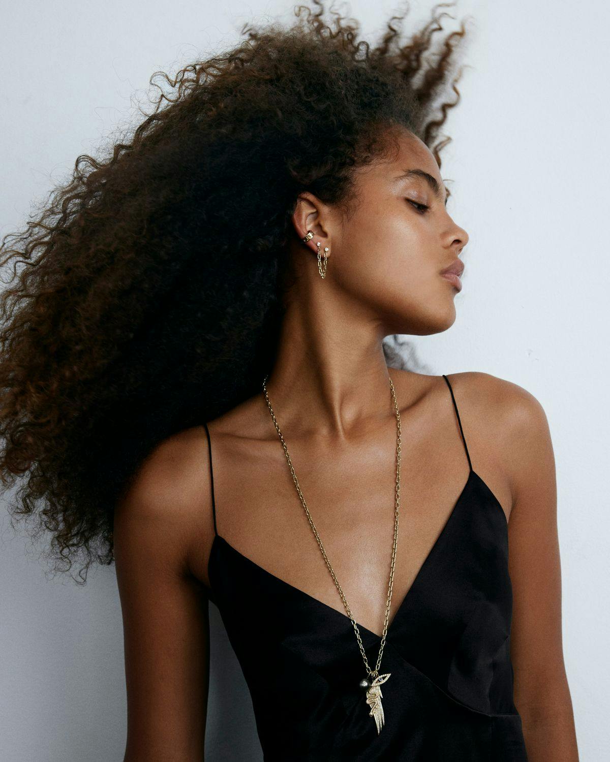 Model with natural hair wearing long chain with assorted charms over black camisole. 