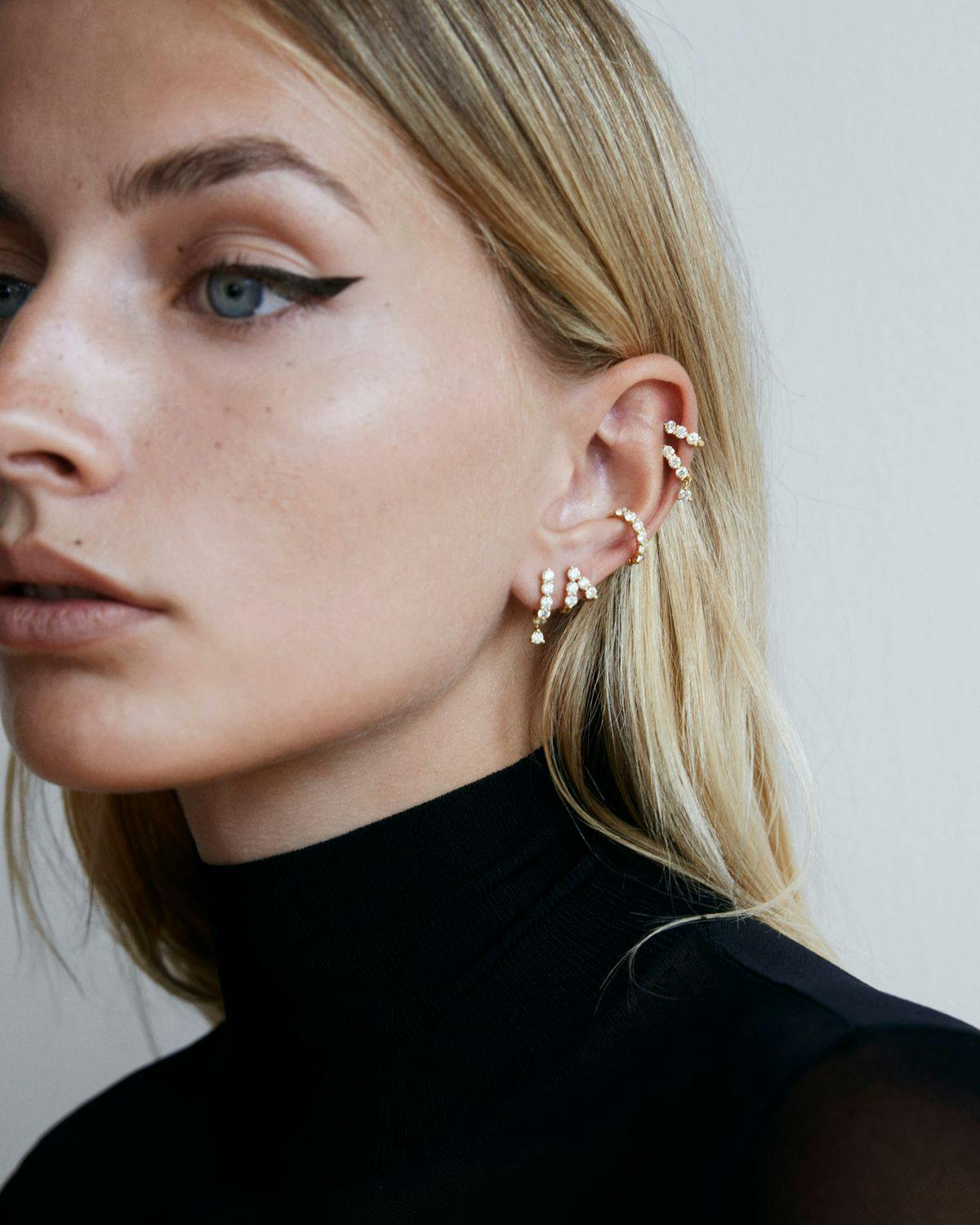 Close up of model looking past camera wearing multiple diamond earrings and earring Huggies on a single ear. 